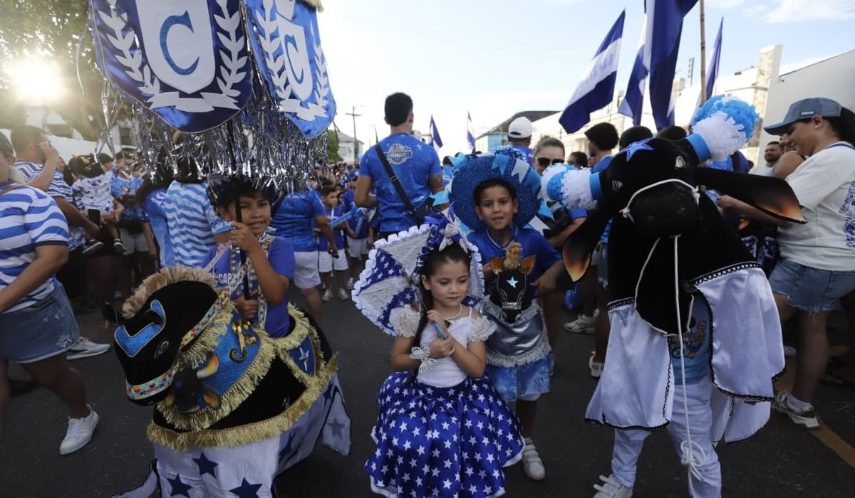 Criança vestida com indumentária azul e branca puxa corda de boi de madeira em rua asfaltada de Parintins.
