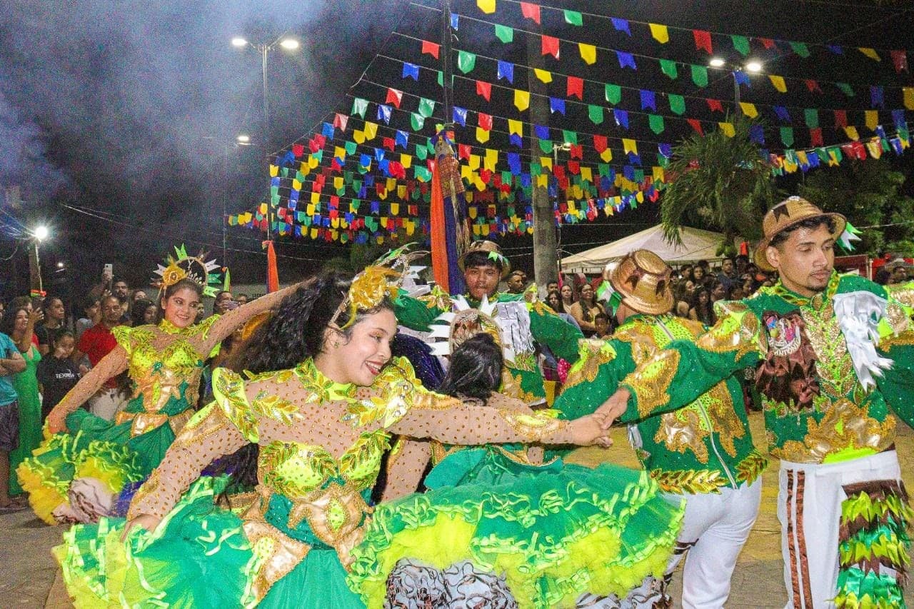 Dançarinos de quadrilha junina com vestidos coloridos e brilhosos se apresentam em palco montado em calçadão.