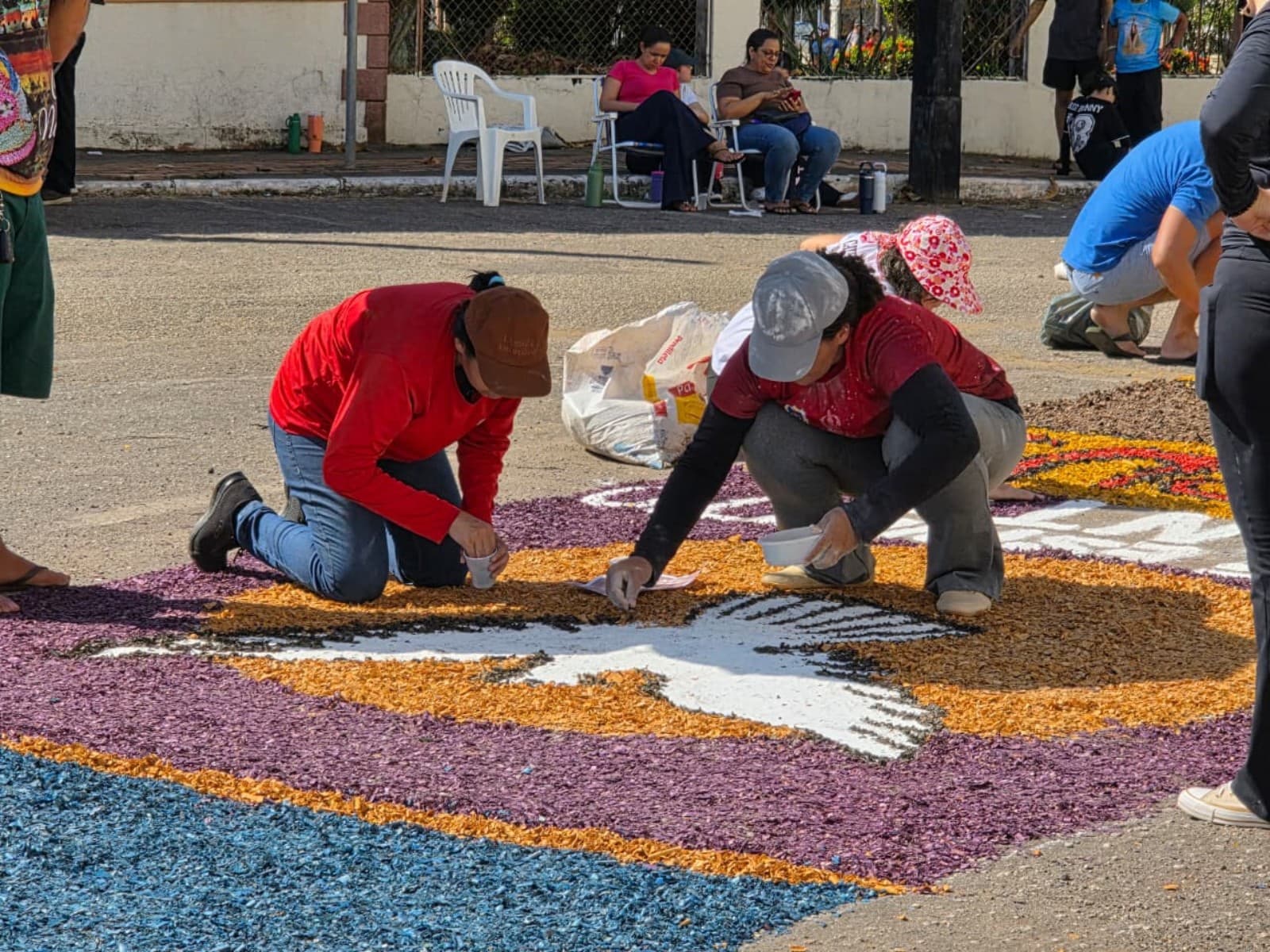 Mãos espalham serragem colorida no chão de rua formando desenhos religiosos em frente a uma catedral.